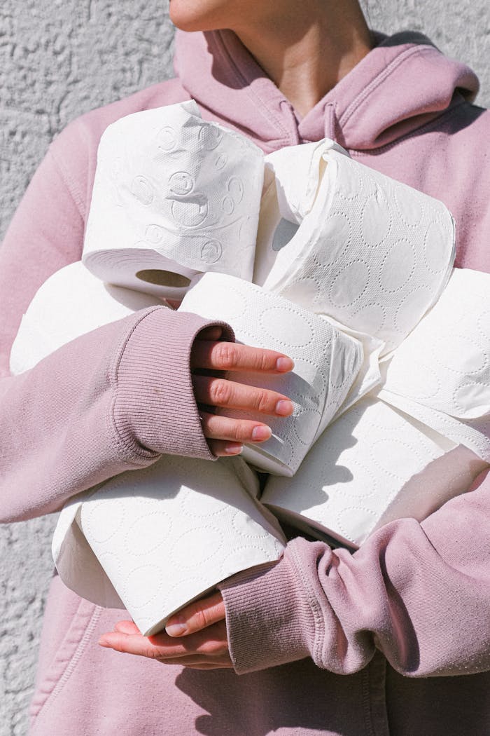 Person in a pink hoodie holding several toilet paper rolls outside against a textured wall.