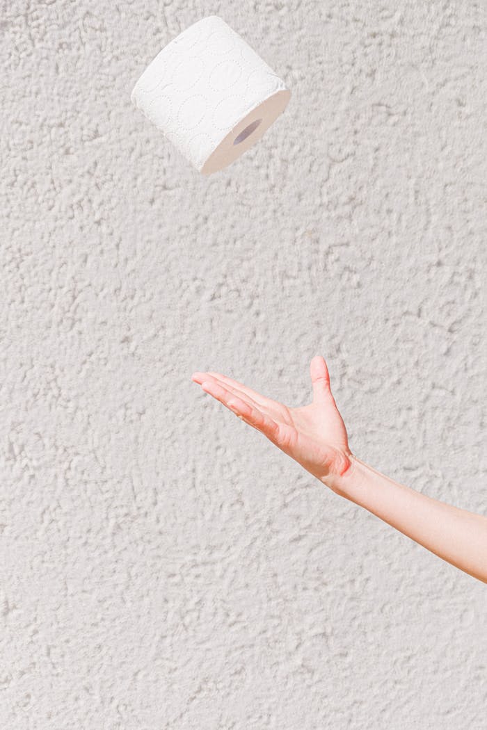 A roll of toilet paper suspended mid-air above an open hand against a textured wall.